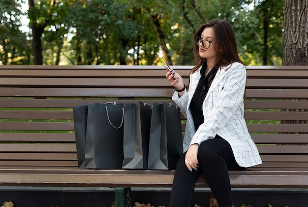 Comprador de mujer se sienta en un banco junto a bolsas de la compra y sostiene el teléfono. Las compras en línea.