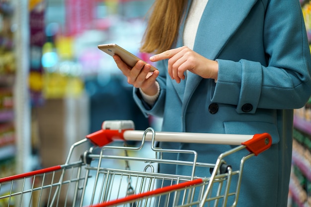 Compradora con carro en el pasillo de la tienda con lista de compras en el teléfono inteligente durante la compra de alimentos