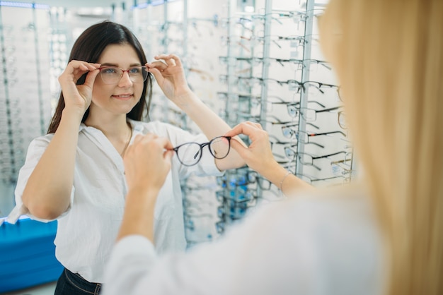 Compradora e oculista feminina escolhe armação de óculos contra vitrine com óculos na loja de ótica. seleção de óculos com optometrista profissional