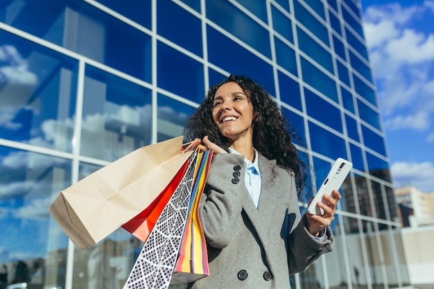 Compradora feliz do lado de fora perto da grande loja de shopping segurando smartphone e pacotes coloridos com mercadorias