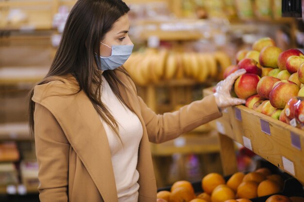 Compradora usando uma máscara protetora e luvas no supermercado. Compras durante a quarentena pandêmica. Mulher comprando frutas frescas