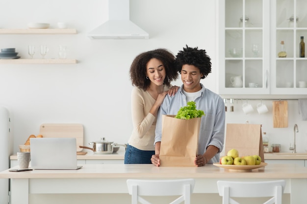 Comprando juntos vegetais para nutrição e culinária adequadas, relacionamentos familiares e tarefas domésticas. Feliz jovem casal afro-americano com saco de comida ecológica no interior da cozinha com laptop, copie o espaço