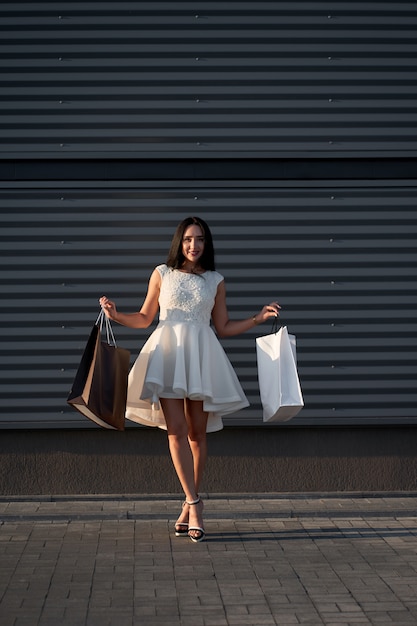 Compras. Atractiva mujer elegante vestido blanco de moda con bolsas de compras en la pared gris
