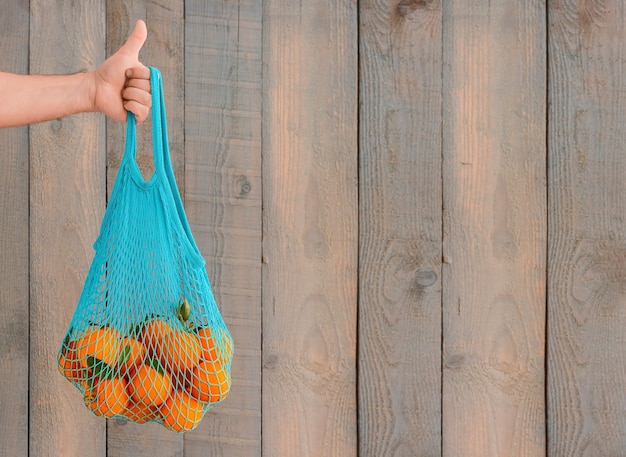 Compras de comestibles sin bolsas de plástico. Concepto de desperdicio cero. La mano de un hombre sostiene una bolsa reutilizable ecológica con frutas orgánicas. Copie el espacio, fondo de madera