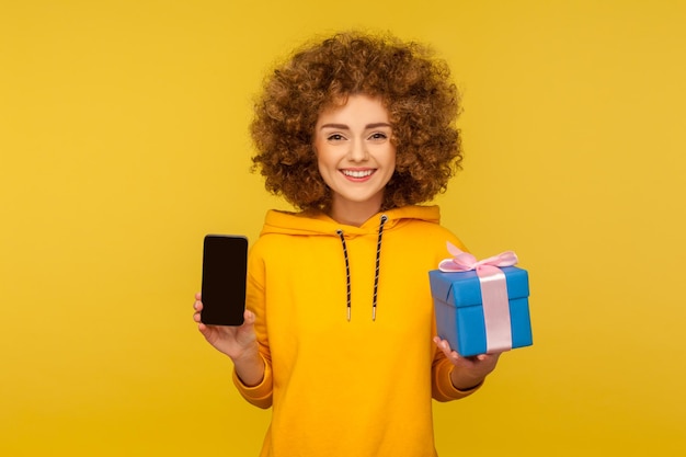 Compras comprando regalos en línea Retrato de una joven feliz de pelo rizado con capucha de estilo urbano sosteniendo un teléfono celular y una caja de regalo sonriendo a la cámara en un estudio interior aislado en un fondo amarillo