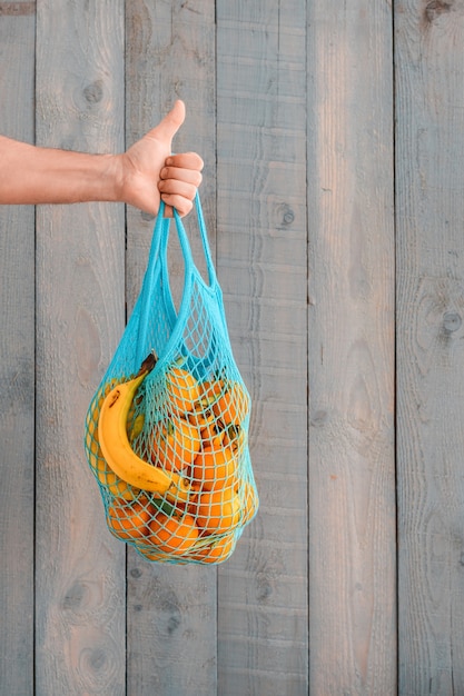 Compras de mantimentos sem sacolas plásticas. Conceito de desperdício zero. Uma mão masculina segura uma sacola reutilizável ecológica com frutas orgânicas, laranjas e bananas. Copiar espaço, parede de madeira