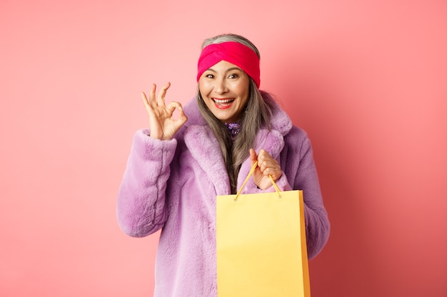 Compras e moda. Feliz mulher asiática sênior em roupas elegantes, segurando o saco de papel da loja, mostrando o sinal de OK, recomendando a loja, fundo rosa.