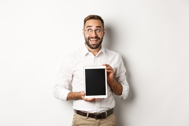 Compras e tecnologia. Homem pensativo mostrando a tela do tablet digital, olhando para o canto superior esquerdo e pensando, em pé sobre um fundo branco.