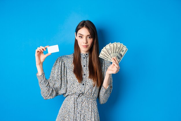 Compras. Elegante mujer descarada con billetes de un dólar dinero y tarjeta de crédito de plástico, mirando confiado en la cámara, azul.