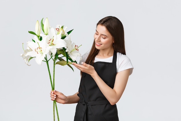 Compras, empleados y concepto de pequeña empresa. Floristería tierna linda en la tienda de flores haciendo un ramo perfecto de lirios blancos, tocando la hoja y sonriendo feliz, procesando pedidos en línea, entregando flores.