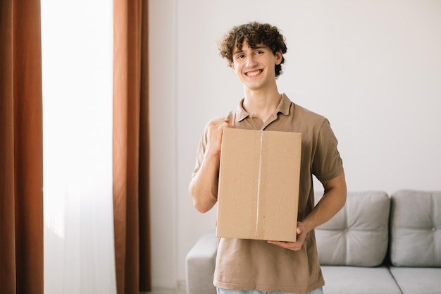 Compras y entrega Rizado joven cliente sosteniendo caja de cartón posando sonriendo a la cámara de pie en casa emocionado comprador masculino abrazando paquete entregado concepto de adicción a las compras