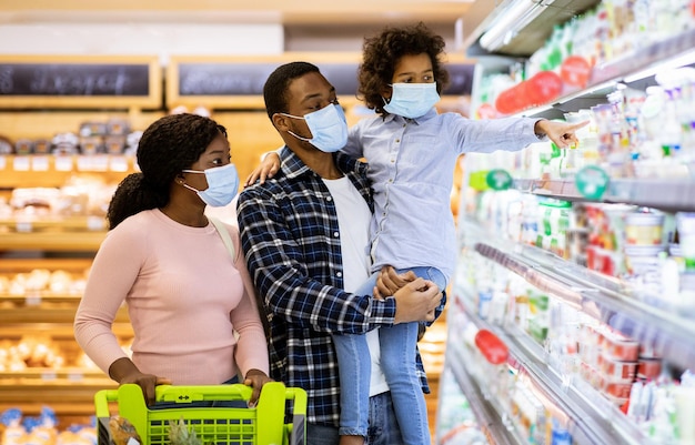 Compras familiares durante la pandemia de coronavirus. Familia afroamericana con niños con mascarillas, comprando alimentos en el supermercado, panorama. Padres negros con sus hijos comprando productos en el centro comercial