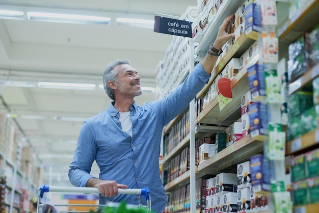 Compras felices y hombre en el supermercado con estantería para comestibles y productos en la tienda de conveniencia Consumidor minorista y persona con carrito en el pasillo para opciones de elección y compra con descuento
