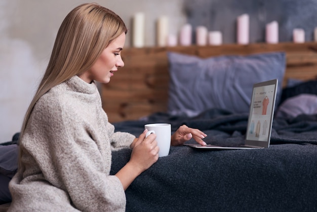 Compras felices. Mujer sonriente bastante joven que tiene compras en línea y que usa la computadora portátil mientras toma café.