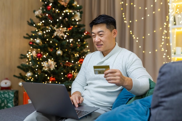 Compras festivas en línea, un joven asiático está sentado en el sofá cerca del árbol de navidad en casa.