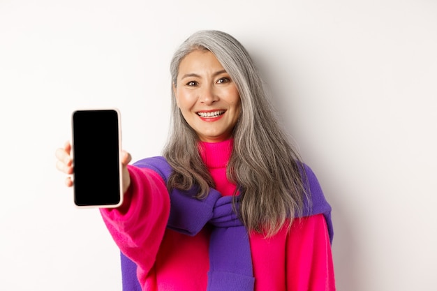 Las compras en línea. Cerca de la elegante mujer mayor asiática extendiendo la mano con el teléfono móvil, mostrando la pantalla del smartphone en blanco y sonriendo, de pie sobre fondo blanco.