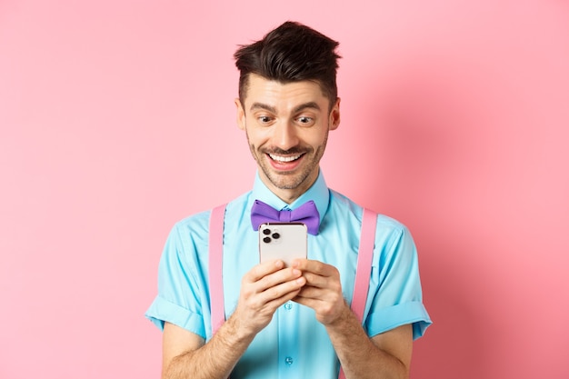 Las compras en línea. Chico feliz mirando la pantalla del teléfono inteligente, leyendo el mensaje y sonriendo, de pie sobre fondo rosa.