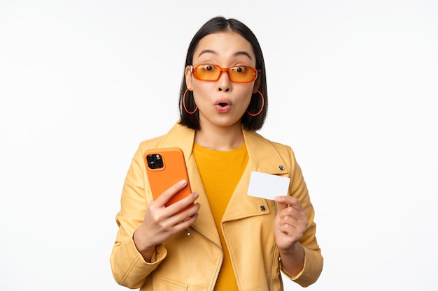 Compras en línea Elegante modelo femenino asiático en gafas de sol con tarjeta de crédito y teléfono móvil sonriendo feliz de pie sobre fondo blanco Copiar espacio