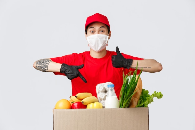 Compras en línea, entrega de alimentos y concepto de pandemia de coronavirus. Repartidor carismático con uniforme rojo, máscara médica y guantes, muestra el pulgar hacia arriba y la aprobación, apuntando al paquete de comestibles