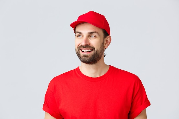 Compras en línea, entrega durante la cuarentena y concepto de comida para llevar. Chico barbudo alegre con gorra de uniforme rojo y camiseta, mirando a otro lado con sonrisa complacida, leyendo letrero de banner, fondo gris