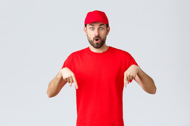 Compras en línea, entrega durante la cuarentena y concepto de comida para llevar. El mensajero sorprendido e intrigado informa a los clientes, el empleado con gorra de uniforme rojo y camiseta apuntando con el dedo hacia abajo, dice wow