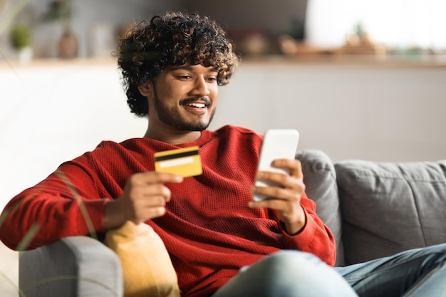 Compras en línea hombre indio sonriente usando teléfono inteligente y tarjeta de crédito en casa