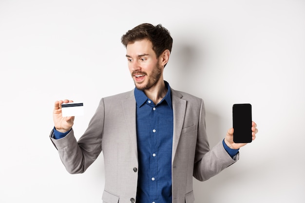 Las compras en línea. Hombre de negocios sonriente en traje que muestra la tarjeta de crédito plástica con la pantalla vacía del teléfono inteligente, de pie contra el fondo blanco.