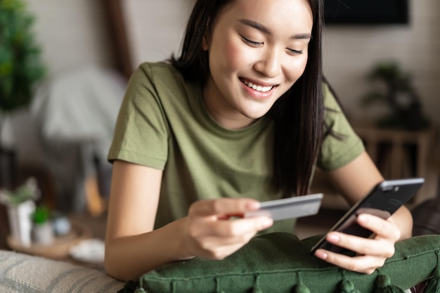 Compras en línea joven mujer asiática sentada en casa y hacer compras en la aplicación de teléfono móvil sosteniendo cr ...