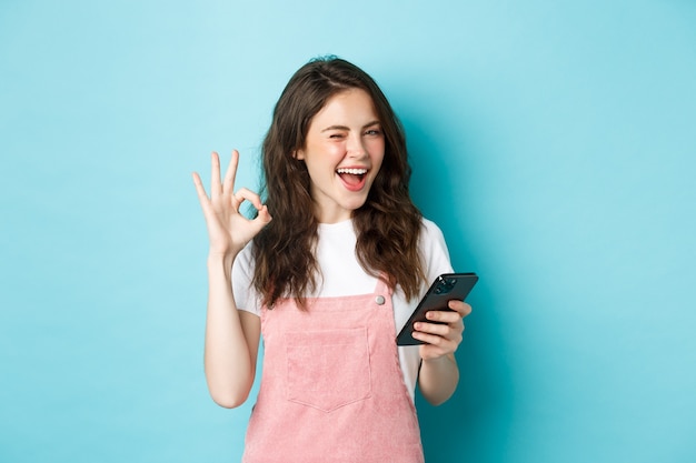 Las compras en línea. Linda chica alegre guiñando un ojo, sonriendo y mostrando un signo de bien después de usar la aplicación de teléfono inteligente, recomendando una tienda de Internet o una página de redes sociales, fondo azul.