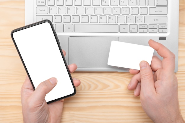 Las compras en línea. Manos sostienen el teléfono celular con pantalla en blanco en blanco y tarjeta de crédito en la computadora portátil