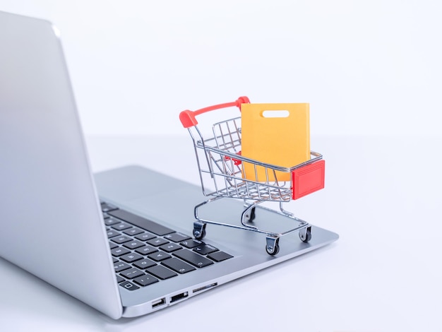 Compras en línea Mini carrito de la tienda con bolsas de papel coloridas sobre una computadora portátil en fondo de mesa blanca comprar en el concepto de casa de cerca