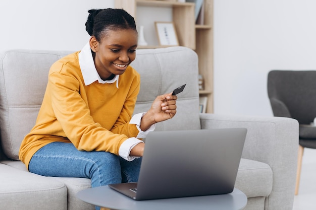 Las compras en línea. Niña negra positiva que usa una computadora portátil y una tarjeta de crédito en casa, hace compras en Internet, se sienta en el sofá en la sala de estar