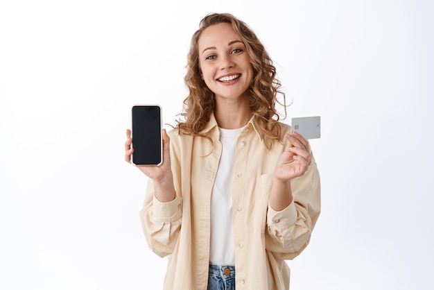 Compras en línea y tecnología Una mujer rubia sonriente muestra una pantalla de teléfono móvil vacía con una tarjeta de crédito de plástico que se ve satisfecha recomienda una aplicación de fondo blanco