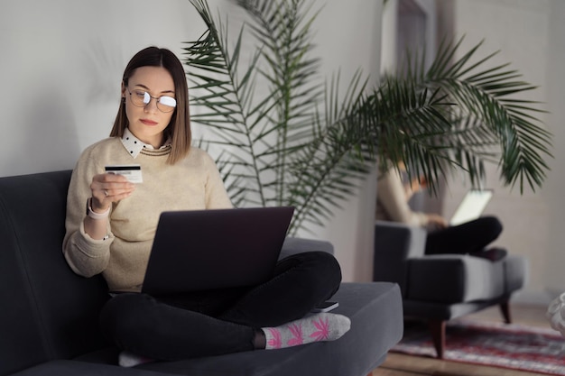 Compras en línea usando tarjeta y computadora portátil Mujer caucásica usando tecnología para comprar mientras está sentada en un sofá en el interior