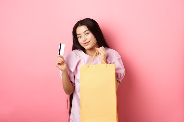 Compras. Menina asiática elegante mostrando a sacola e o cartão de crédito de plástico, de pé no fundo rosa.