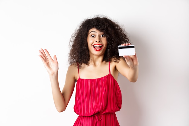 Compras. Mulher de cabelos encaracolados espantada com um vestido vermelho, mostrando o cartão de crédito de plástico e grito de emoção, de pé sobre um fundo branco.