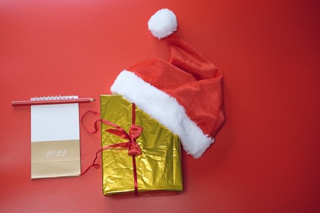 Compras de Navidad. Lista de compras y regalos para el nuevo año. Sombrero de Papá Noel se encuentra en una caja de oro sobre un fondo rojo.