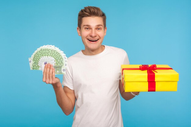 Compras navideñas, bonificaciones. Feliz hombre sonriente alegre con camiseta blanca con caja de regalo y billetes en euros, listo para comprar regalos y recibir reembolso. Foto de estudio interior aislado sobre fondo azul.