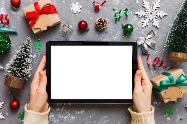 Compras navideñas en línea desde casa manos femeninas sosteniendo una tableta con pantalla blanca en blanco vista superior mano de mujer sosteniendo una tableta con pantalla en blanco Árbol de Navidad y regalos en el fondo