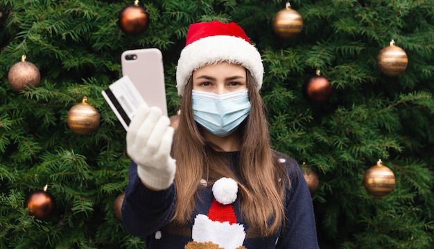 Compras navideñas online. Close up Retrato de mujer con un sombrero de santa claus y una máscara médica con emoción. En el contexto de un árbol de Navidad. Pandemia de coronavirus