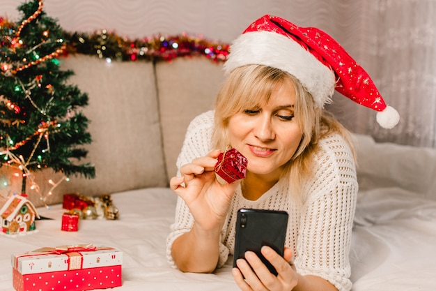 Compras navideñas online. Compradora femenina hace un pedido en el teléfono móvil. Mujer comprar regalos, prepararse para Navidad, caja de regalo en mano. Rebajas de vacaciones de invierno.