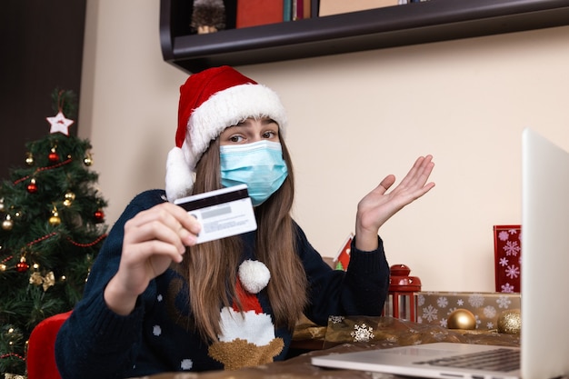 Compras navideñas online. Una niña con un sombrero de santa claus y un suéter azul con una máscara médica se sienta cerca de una computadora portátil. La habitación está decorada de forma festiva. Navidad durante el coronavirus