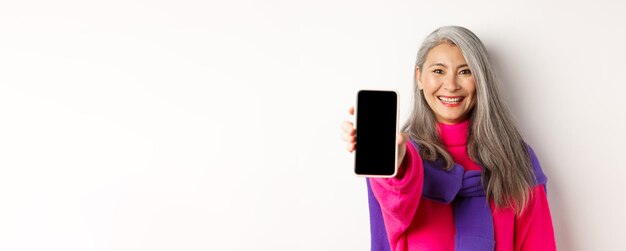 Compras on-line de uma mulher asiática moderna sênior estendendo a mão com o celular mostrando em branco