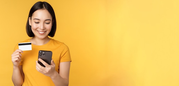 Compras on-line Menina asiática sorridente usando cartão de crédito e aplicativo de celular pagando pedido sem contato no aplicativo de smartphone sobre fundo amarelo