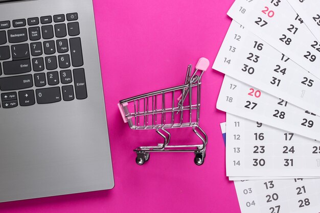 Compras online. Carrinho de supermercado com laptop, calendário mensal em uma superfície rosa. Vista do topo