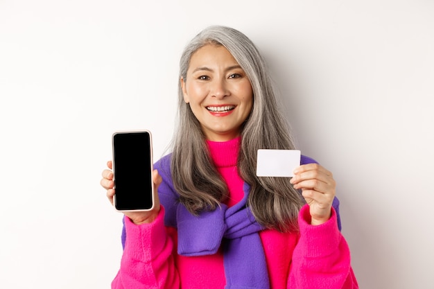 Compras online. Close de uma mulher asiática sênior sorridente, mostrando a tela do celular em branco e um cartão de crédito de plástico, parecendo feliz, em pé sobre um fundo branco