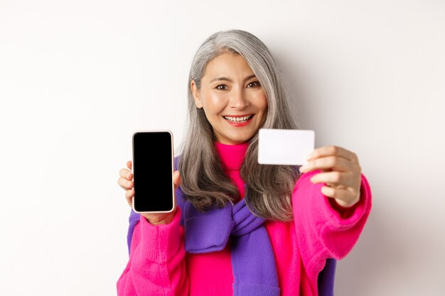 Compras online. Close-up de elegante mulher coreana sênior mostrando a tela do celular vazia e cartão de crédito de plástico, sorrindo feliz, em pé sobre um fundo branco.