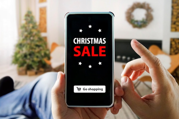 Compras online de natal com telefone. homem senta em um apartamento e faz compras por meio de um smartphone em uma loja online. árvore de natal, presentes, luzes e decorações em segundo plano.