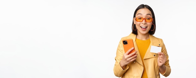 Compras online elegante modelo feminino asiático em óculos de sol segurando cartão de crédito e celular sorrindo feliz em pé sobre fundo branco copie o espaço