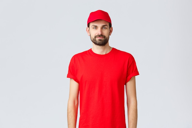 Foto compras online, entrega durante a quarentena e conceito de take-away. jovem mensageiro barbudo em uniforme vermelho, funcionário de boné e camiseta, olhando para a câmera, esperando para anotar seu pedido
